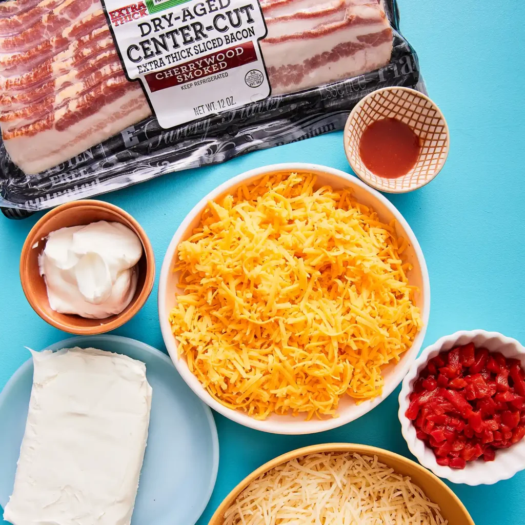 Overhead flat lay of baked pimento dip ingredients including a package of Jones Dairy Farm Dry Aged Cherrywood Smoked Bacon, shredded sharp cheddar, Parmesan, cream cheese, sour cream, diced pimentos, and hot sauce on a teal surface.
