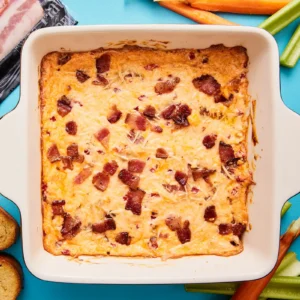Overhead view of a finished baked pimento dip with Jones Dry Aged Bacon, golden melted cheddar and Parmesan, and crispy bacon pieces on top, in a white square baking dish surrounded by crostini, carrots, and celery for dipping.