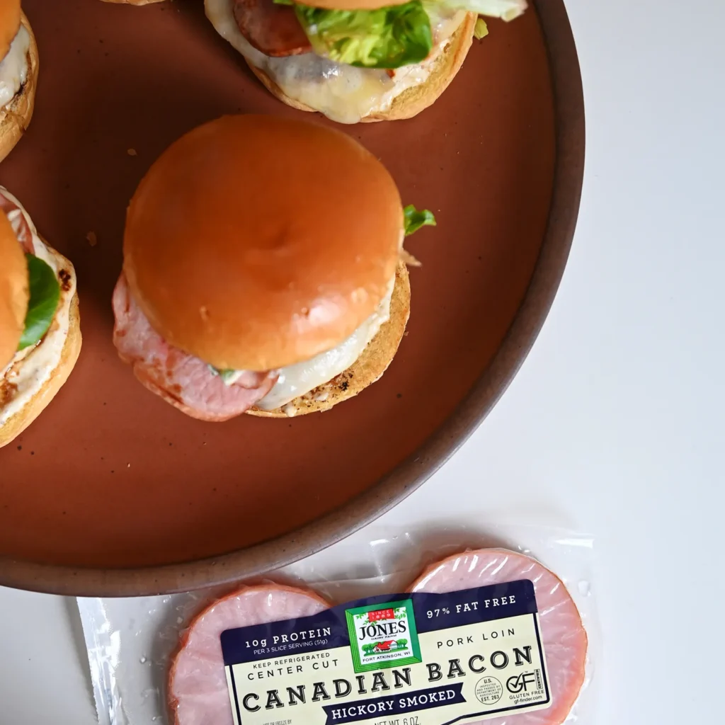 verhead view of chicken cordon bleu burgers on a brown plate alongside a package of Jones Dairy Farm Canadian Bacon Hickory Smoked, showing the Swiss cheese, Canadian bacon, and mixed greens assembly.