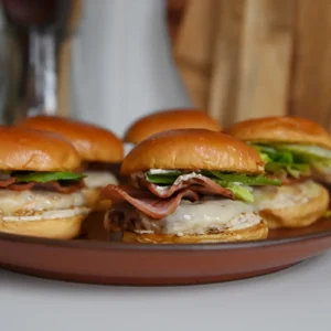 Three chicken cordon bleu burgers on a brown plate, each stacked with a Parmesan and Dijon ground chicken patty, melted Swiss cheese, crisped Jones Canadian Bacon, and mixed greens on a toasted brioche bun.