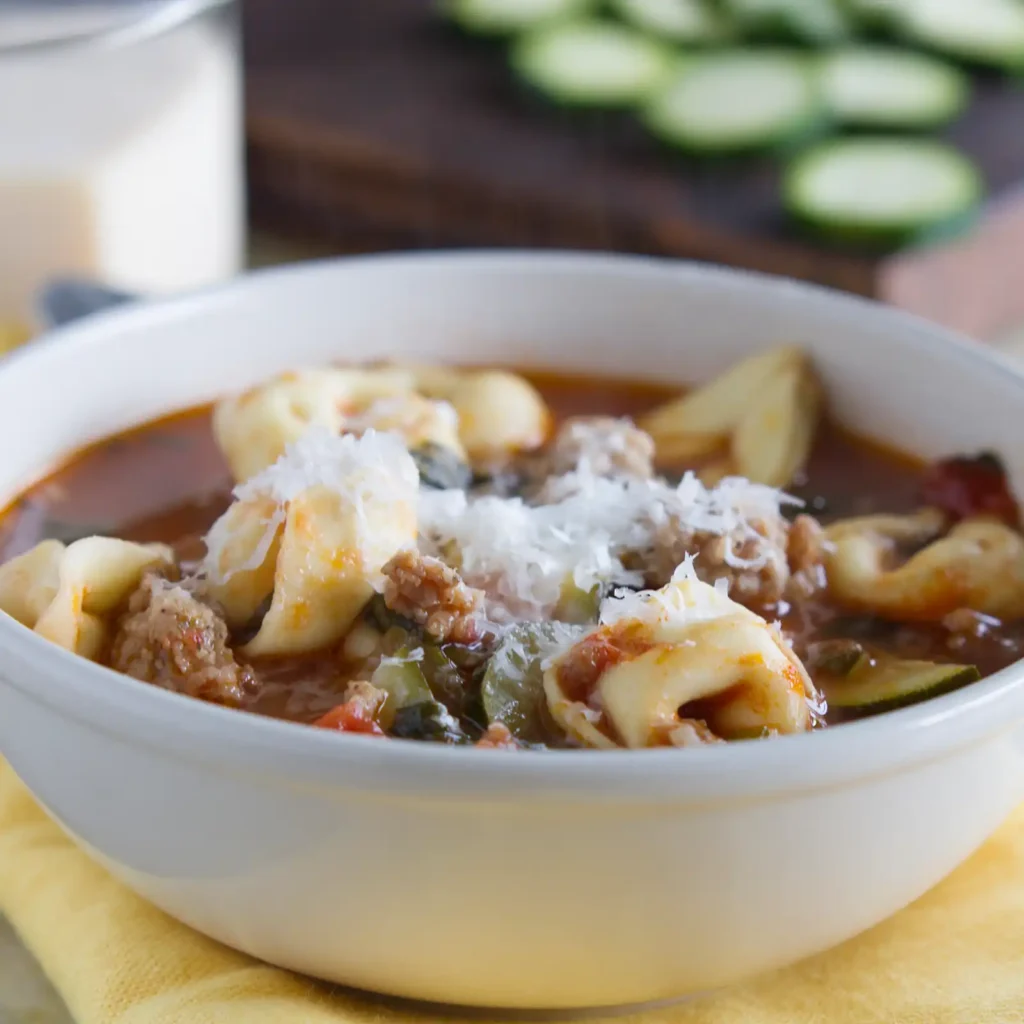 Close-up of Jones sausage and tortellini soup in a white bowl, filled with cheese tortellini, crumbled sausage, zucchini, and tomato broth, topped with grated parmesan.