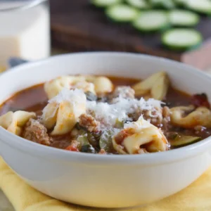 Close-up of Jones sausage and tortellini soup in a white bowl, filled with cheese tortellini, crumbled sausage, zucchini, and tomato broth, topped with grated parmesan.