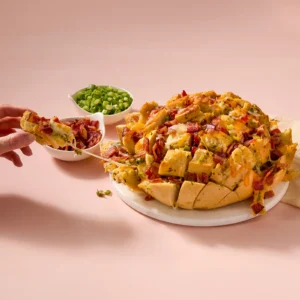 A hand pulling apart a cheesy pull-apart bread loaded with Jones Dairy Farm Dry Aged Cherrywood Smoked Bacon, melted mozzarella and cheddar, green onions, and garlic herb butter, served on a white plate with small bowls of toppings in the background.