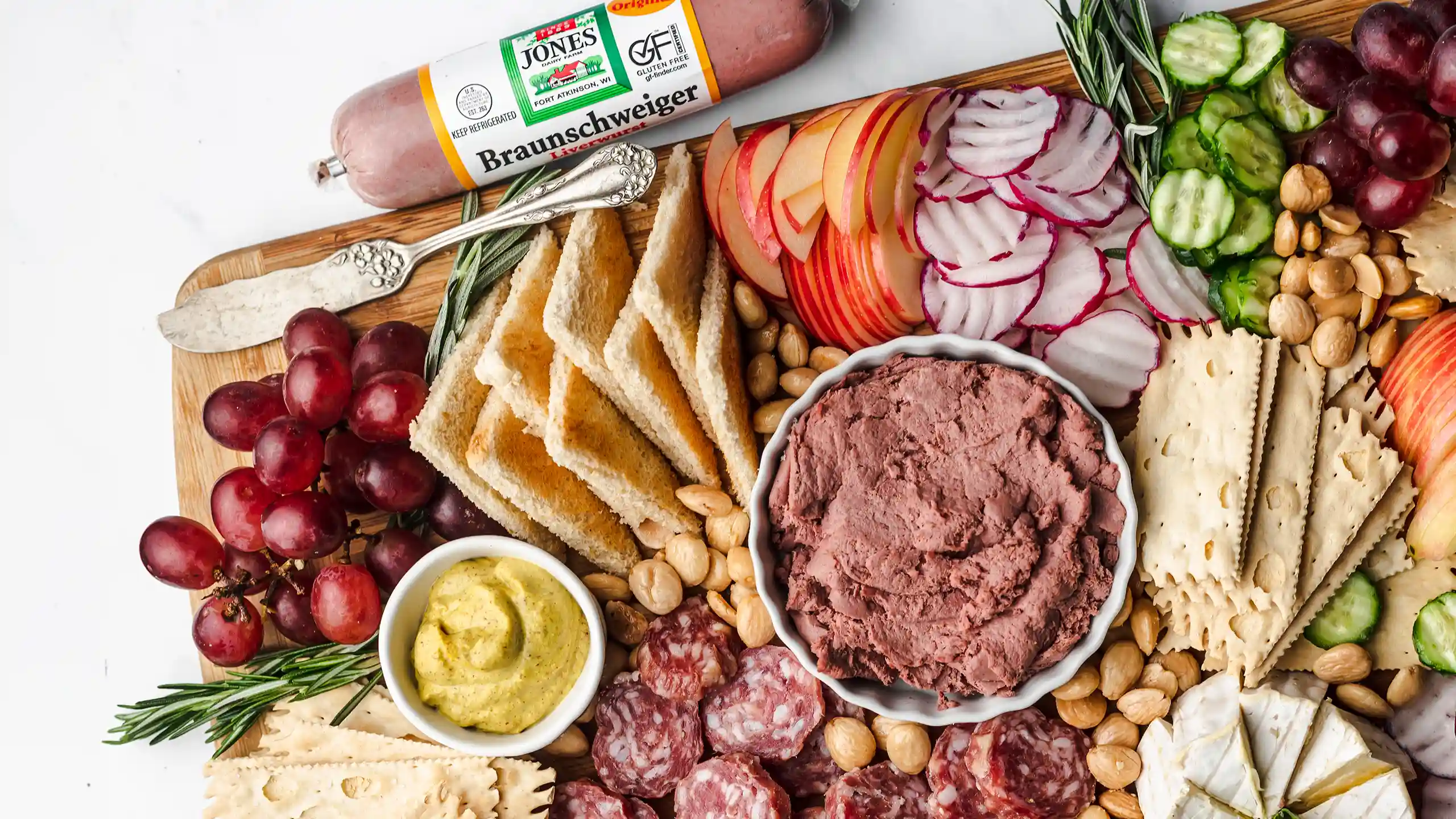 Jones Braunschweiger chub and spread served on charcuterie boards with grapes, toast, crackers, mustard, salami, almonds, apples, radishes, and cucumber