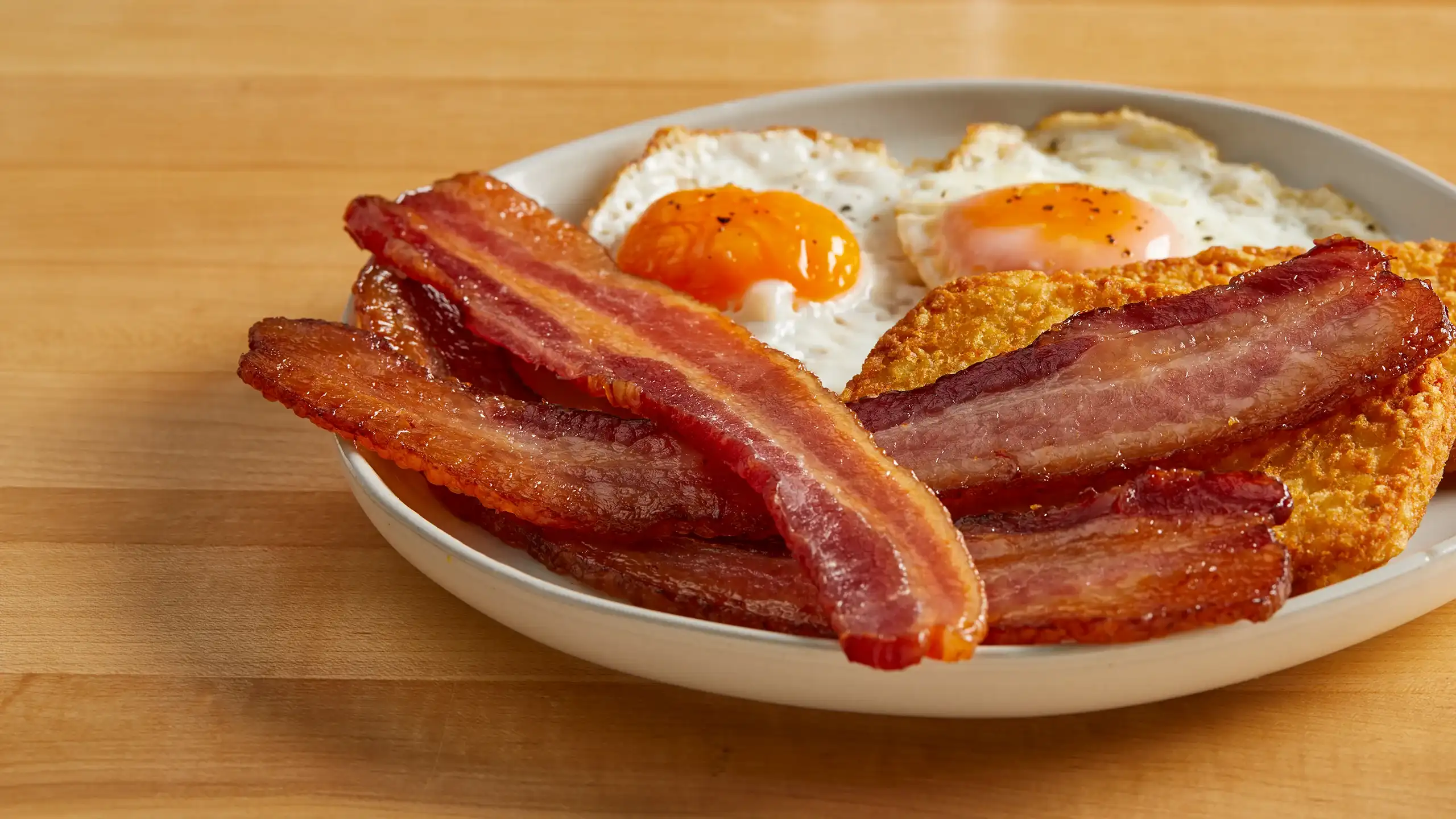 Plate of thick cut bacon, fried eggs, and hashbrowns on wood countertop