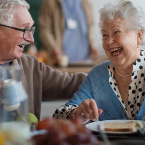 Two seniors laughing together during a meal in a healthcare dining setting
