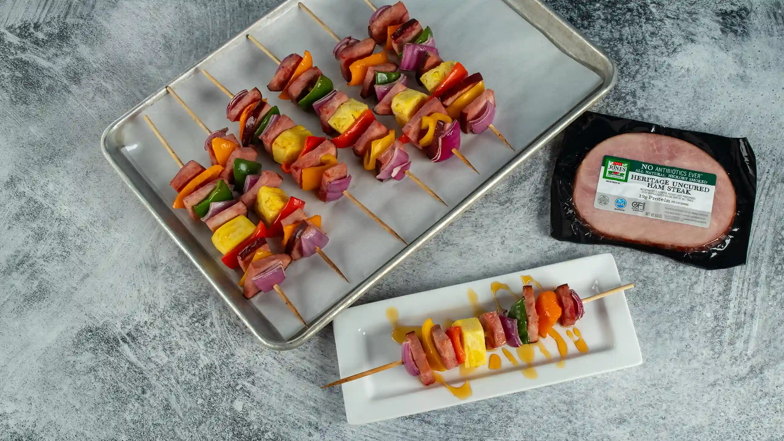 Jones No Antibiotics Ever Heritage Uncured Ham Steak kebabs with peppers, red onion, and pineapple on a sheet pan, with a finished kebab on a serving plate and the product package