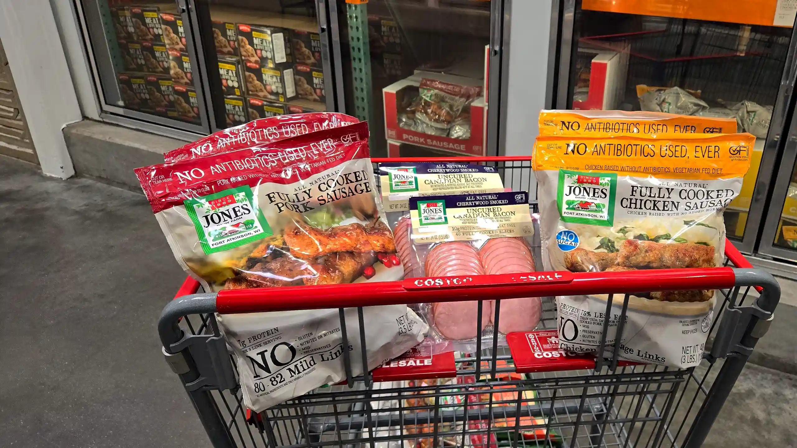 A Costco shopping cart filled with Jones bulk bags of fully cooked pork sausage, fully cooked chicken sausage, and uncured Canadian bacon at a Costco warehouse