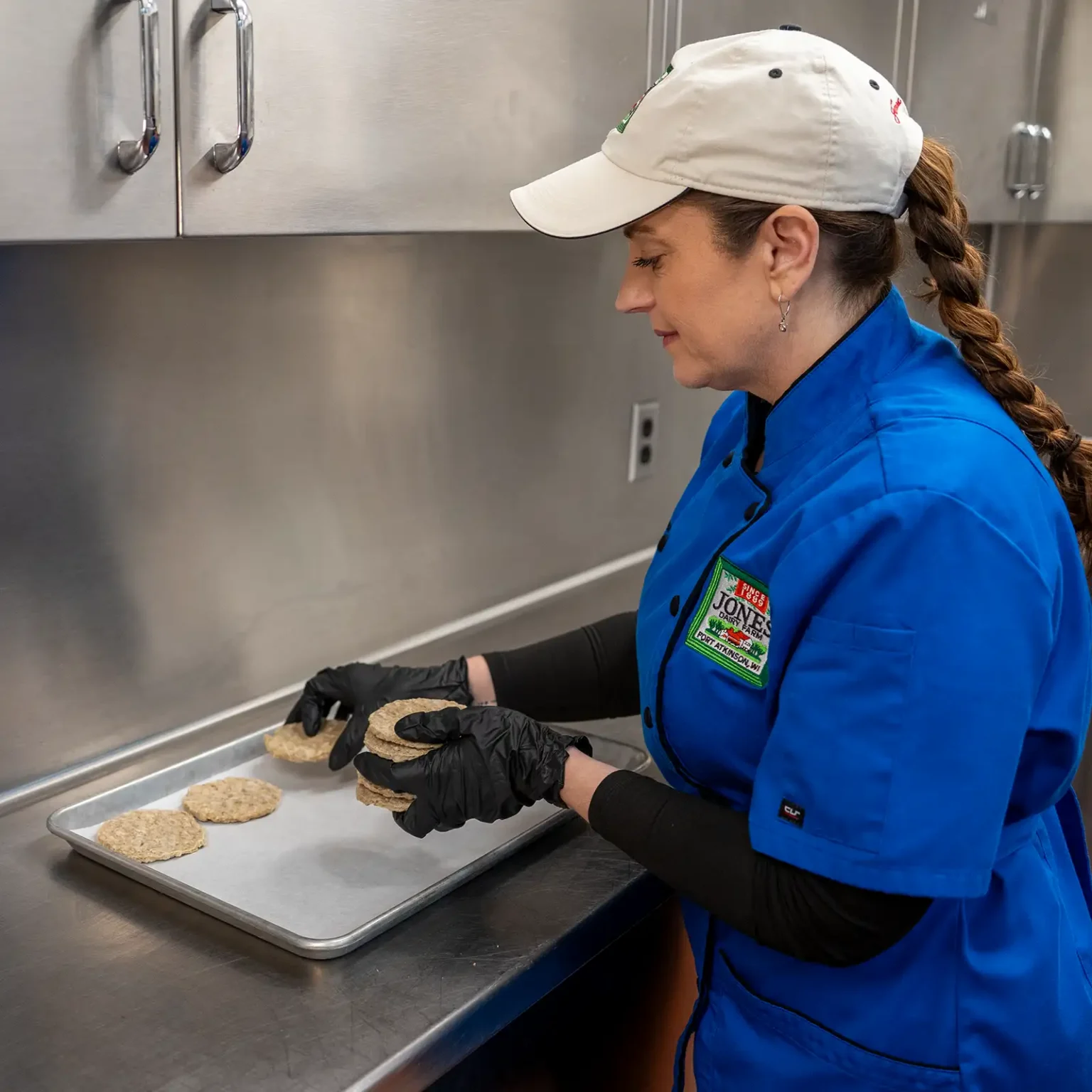 1:59 PMClaude responded: Chef Kate Konen arranges Jones Dairy Farm chicken sausage patties on a parchment-lined sheet pan in a commercial kitchen, wearing a blue chef's coat with a Jon…Chef Kate Konen arranges Jones Dairy Farm chicken sausage patties on a parchment-lined sheet pan in a commercial kitchen, wearing a blue chef's coat with a Jones Dairy Farm logo, black gloves, and a white cap