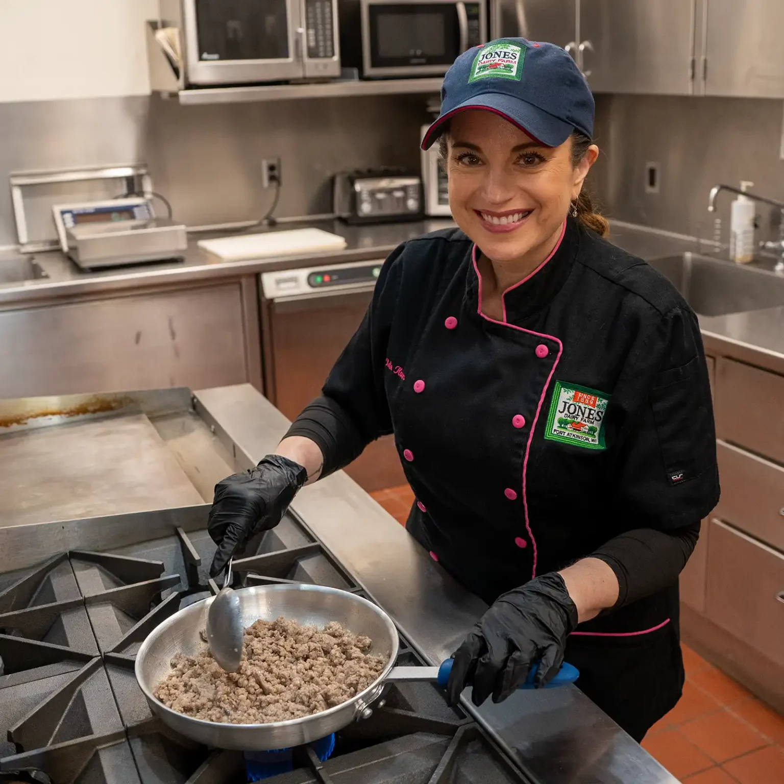 Chef Kate Konen smiles at the camera while cooking Jones Dairy Farm ground sausage in a stainless steel skillet on a commercial kitchen range, wearing a black chef's coat with pink trim and a Jones Dairy Farm logo cap