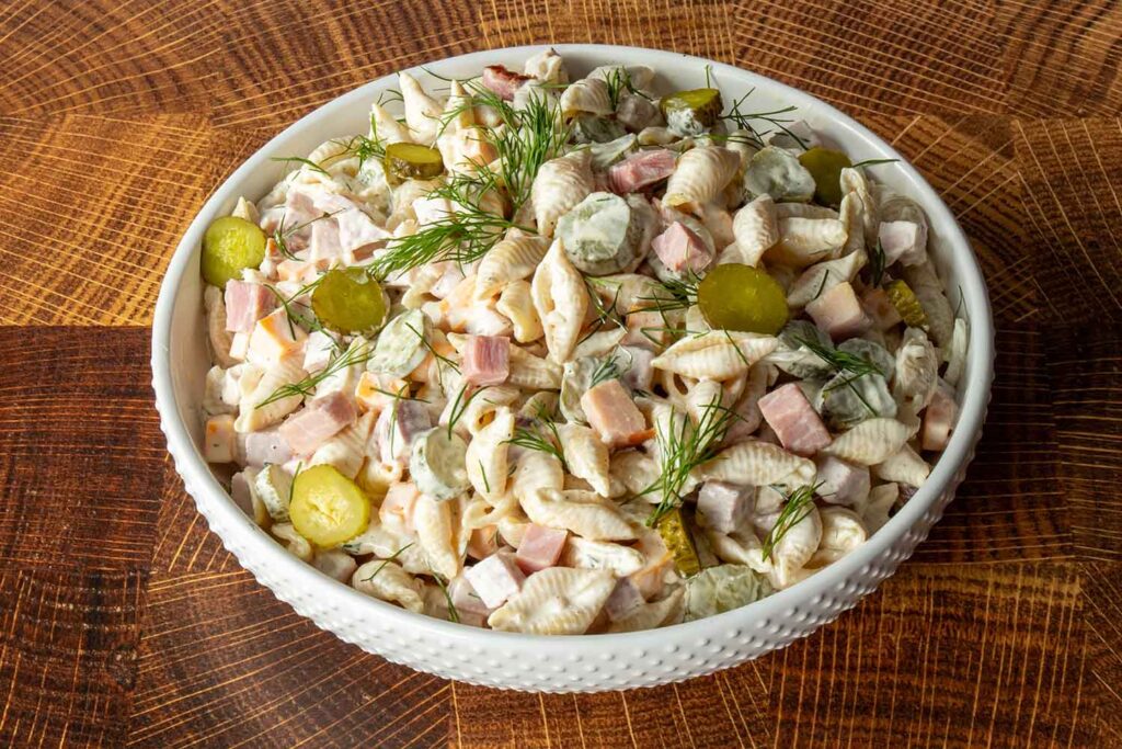 Dill pickle pasta salad with ham in a white hobnail bowl, garnished with fresh dill and pickle coins, on a wood cutting board.