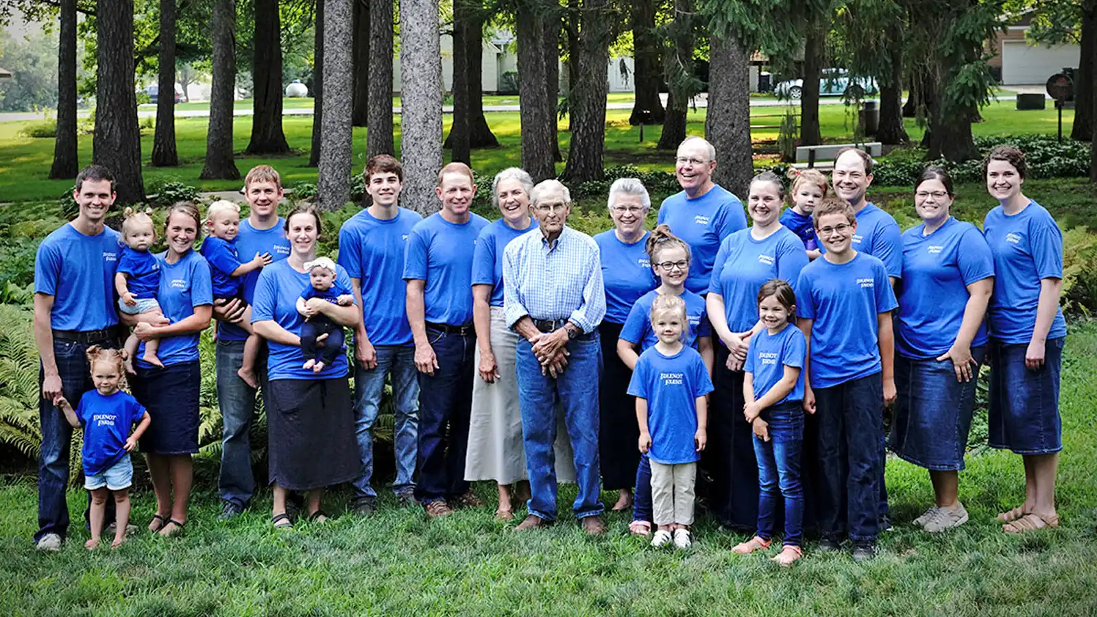 A multigenerational Wholestone Farms family poses outdoors in matching blue Wholestone Farms T-shirts on a grassy lawn surrounded by trees.
