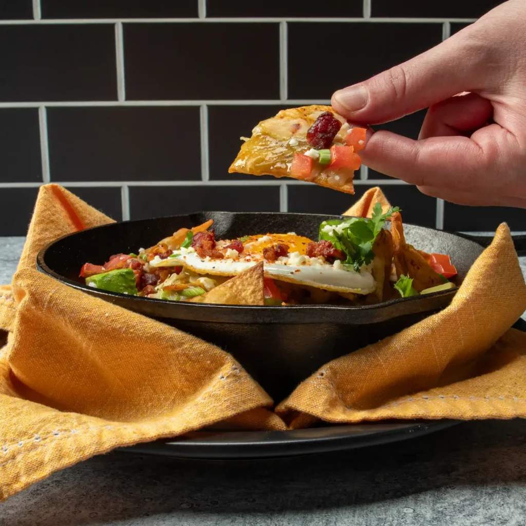 A hand pulls a salsa verde-coated tortilla chip from a cast iron skillet of Bacon Chilaquiles made with Jones Raw Diced Bacon, avocado, queso fresco, and a fried egg