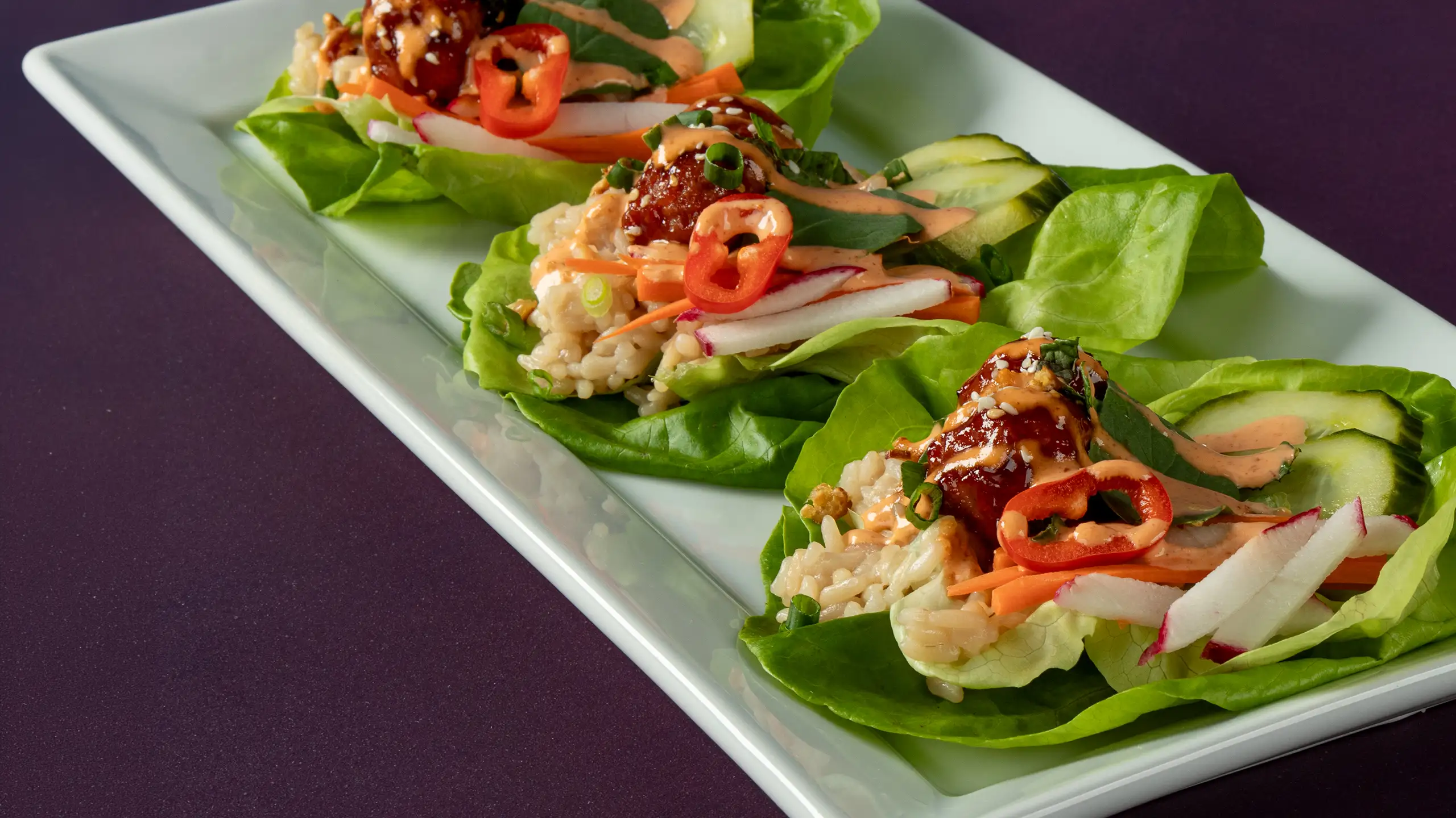 Jones Dairy Farm pork and bacon meatballs in Korean BBQ sauce on Bibb lettuce with sushi rice, pickled radish, shredded carrots, sliced cucumbers, Fresno chilies, and sriracha mayo on a white serving platter