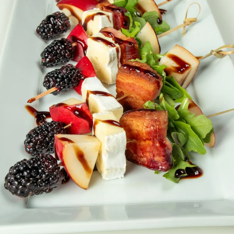 Three skewers on a white rectangular plate, each threaded with fresh blackberries, red Anjou pear, creamy brie, roasted Jones Dry Aged Slab Bacon, and arugula, finished with a blackberry balsamic glaze drizzle.