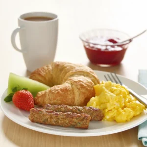 A breakfast plate with Jones Dairy Farm chicken sausage links, scrambled eggs, a croissant, fresh strawberry and honeydew, served alongside a cup of coffee and a small bowl of jam