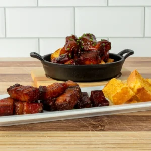 Jones Dairy Farm Cherrywood Smoked Bacon Slab burnt ends glazed in smoky barbecue sauce, served in a cast iron skillet and on a white platter alongside cubed cornbread