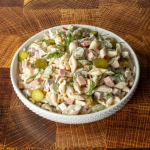 Dill pickle pasta salad with ham in a white hobnail bowl, garnished with fresh dill and pickle coins, on a wood cutting board.
