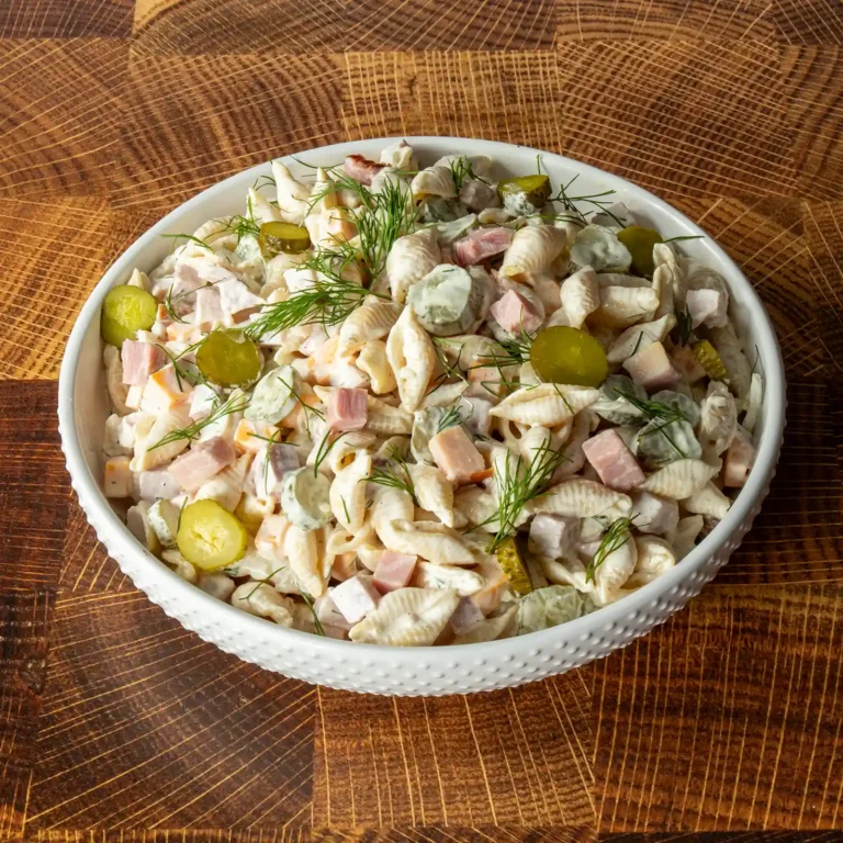 Dill pickle pasta salad with ham in a white hobnail bowl, garnished with fresh dill and pickle coins, on a wood cutting board.