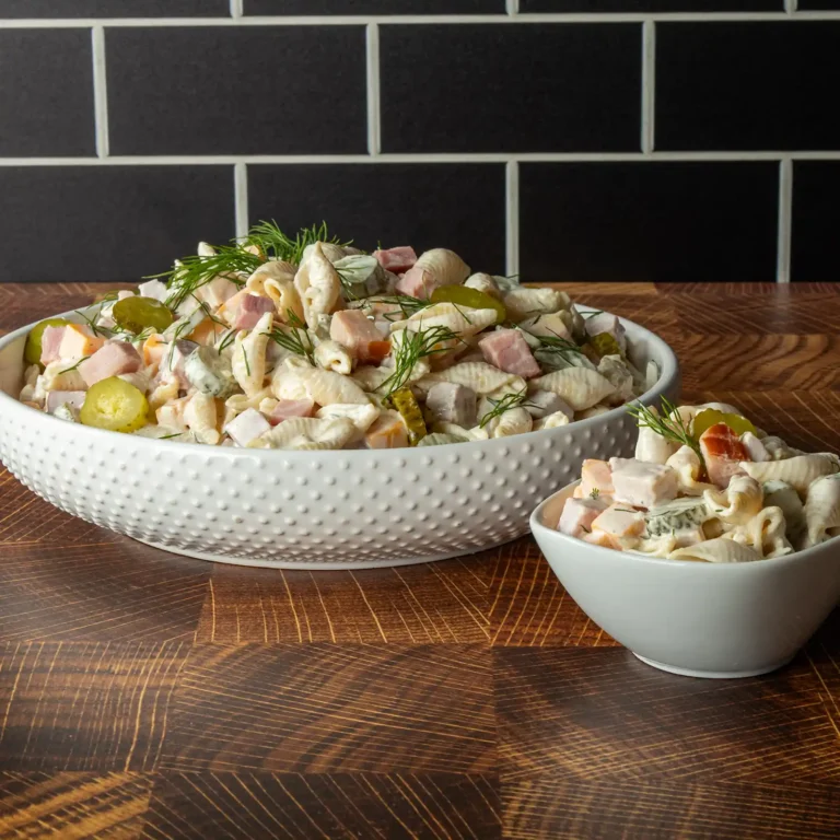 A large serving bowl and individual bowl of dill pickle pasta salad with ham, garnished with fresh dill and pickle coins, on a wood surface with dark tile backdrop.