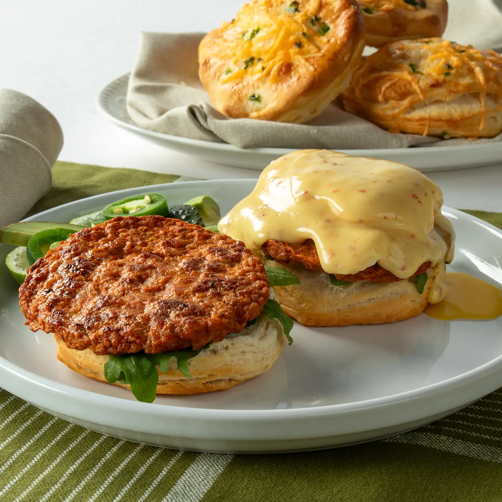 Two open-face jalapeño cheddar biscuits on a white plate, each topped with a Jones Chicken Chorizo Sausage Patty, arugula, and Aleppo pepper Hollandaise, with additional biscuits and sliced jalapeños in the background.