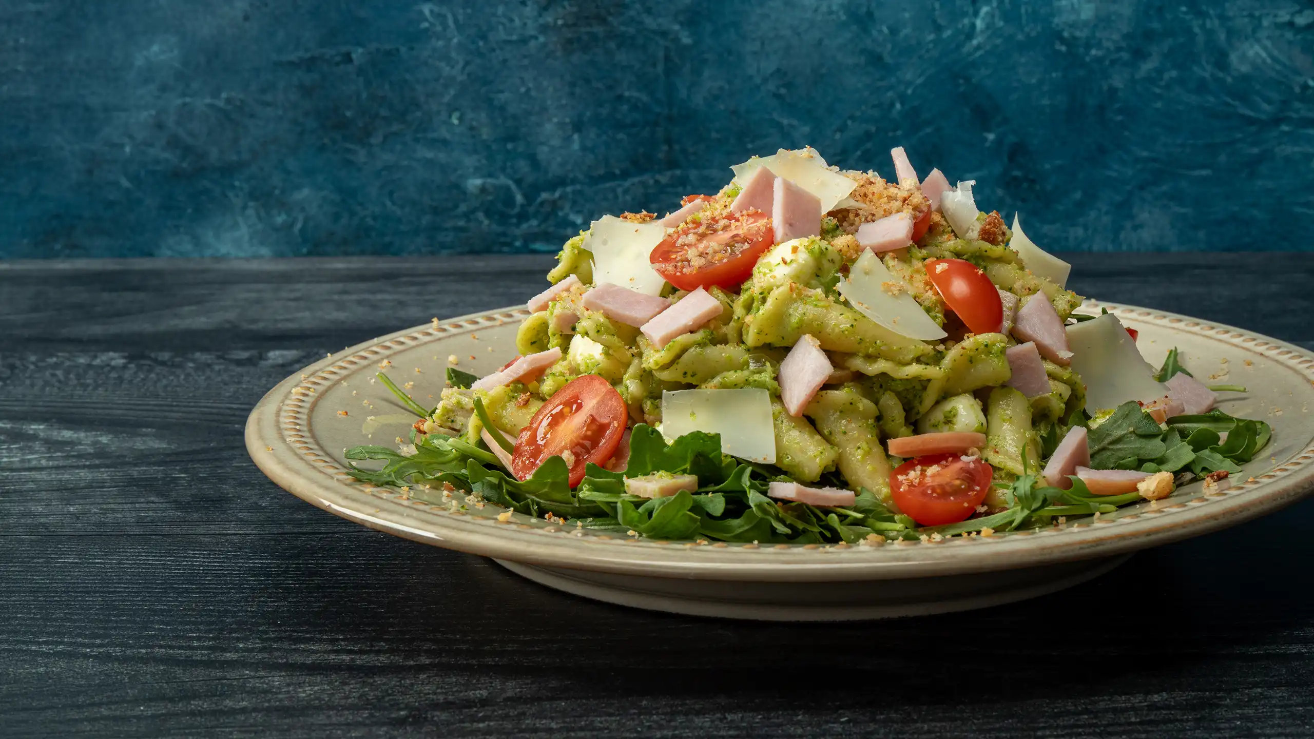 Pesto pasta salad with diced Jones Canadian Bacon, cherry tomatoes, fresh mozzarella, shaved parmesan, and toasted breadcrumbs over arugula