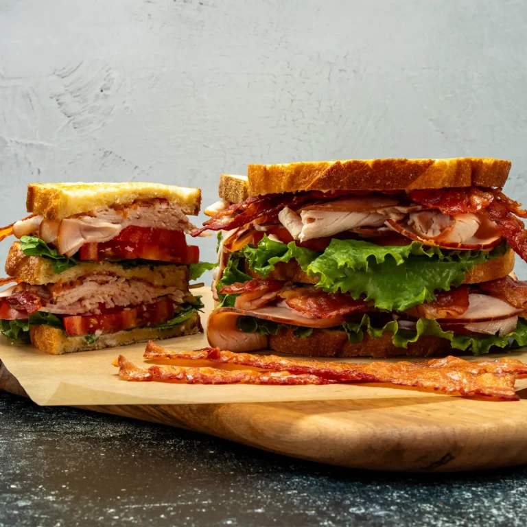 Jones Dairy Farm fully cooked bacon on a triple-decker club sandwich with turkey, ham, lettuce, and tomato on toasted bread, served on a wood cutting board with extra bacon strips alongside.