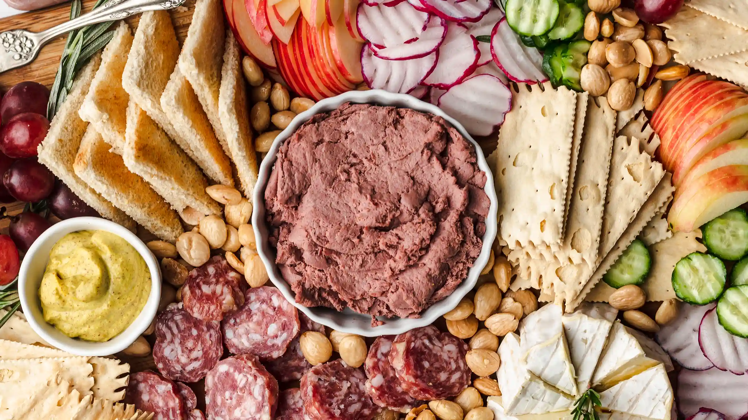 Jones Dairy Farm Braunschweiger spread served on a charcuterie board with sliced salami, brie, mustard, toast points, crackers, apples, radishes, cucumbers, grapes, and marcona almonds