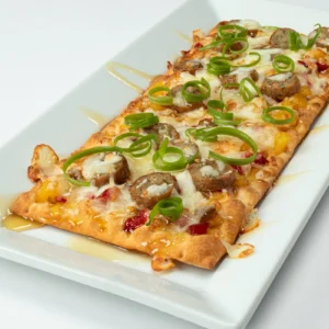 Jones Four Pepper Chicken Sausage flatbread with pineapple-peppadew compote, melted mozzarella, white balsamic drizzle, and curled green onions on a white plate