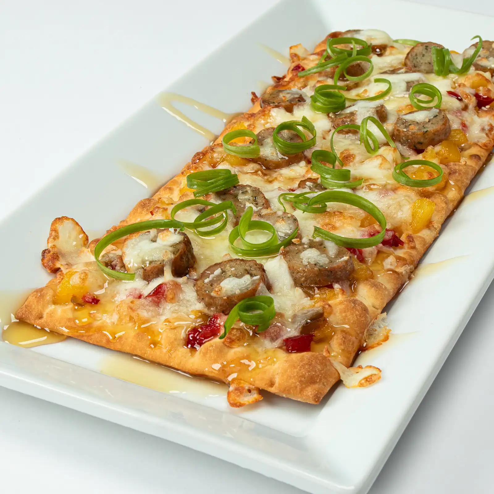 Jones Four Pepper Chicken Sausage flatbread with pineapple-peppadew compote, melted mozzarella, white balsamic drizzle, and curled green onions on a white plate