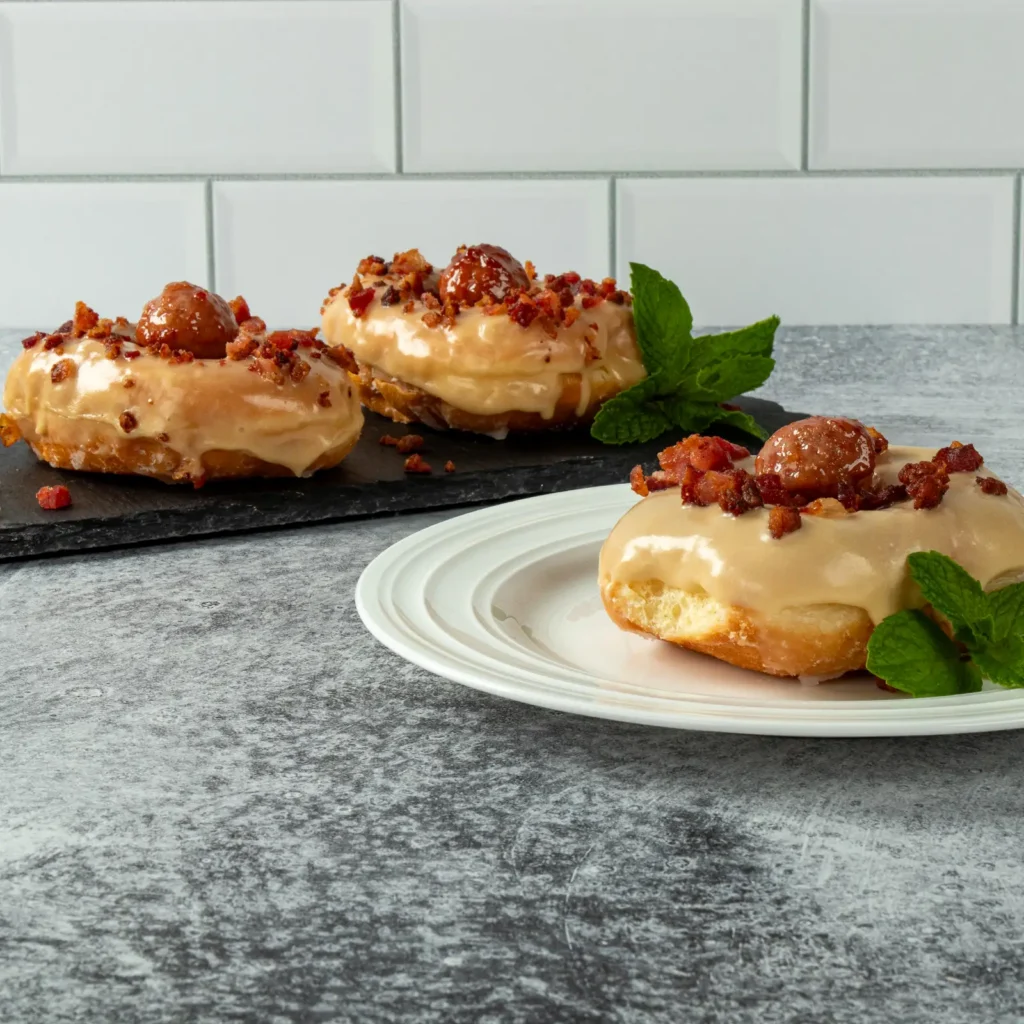 Three maple bacon donuts topped with crispy bacon crumbles and Jones Dairy Farm pork and bacon meatballs displayed on plates and a serving board.