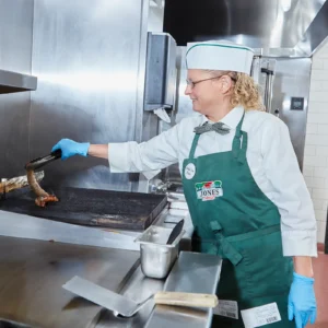 A Jones Market employee named Pam grills sausage links on a commercial grill, wearing a green Jones Market apron, white chef's coat, and blue gloves.
