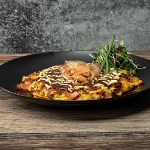A golden-brown okonomiyaki on a black plate, topped with a crosshatch drizzle of okonomiyaki sauce and Kewpie mayonnaise, a mound of bonito flakes, and a cluster of fresh microgreens and chives, with Jones Canadian Bacon visible along the edges of the savory pancake.