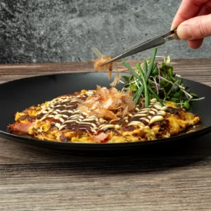 A hand using metal tweezers to place bonito flakes onto a golden-brown okonomiyaki on a black plate, finished with a crosshatch of okonomiyaki sauce and Kewpie mayonnaise, with fresh microgreens and chives arranged alongside Jones Canadian Bacon visible along the edge of the pancake.