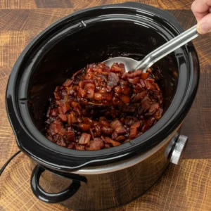 A hand ladling bacon baked beans from a slow cooker on a wood surface.