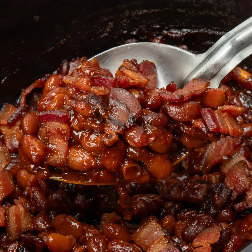 A ladle scooping smoky bacon baked beans from a slow cooker.