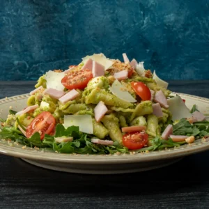 A mound of pasta tossed in bright green broccolini pesto, served over baby arugula and topped with diced Canadian bacon, halved cherry tomatoes, shaved Pecorino Romano, and toasted garlic breadcrumbs on a cream-rimmed plate against a deep blue background.