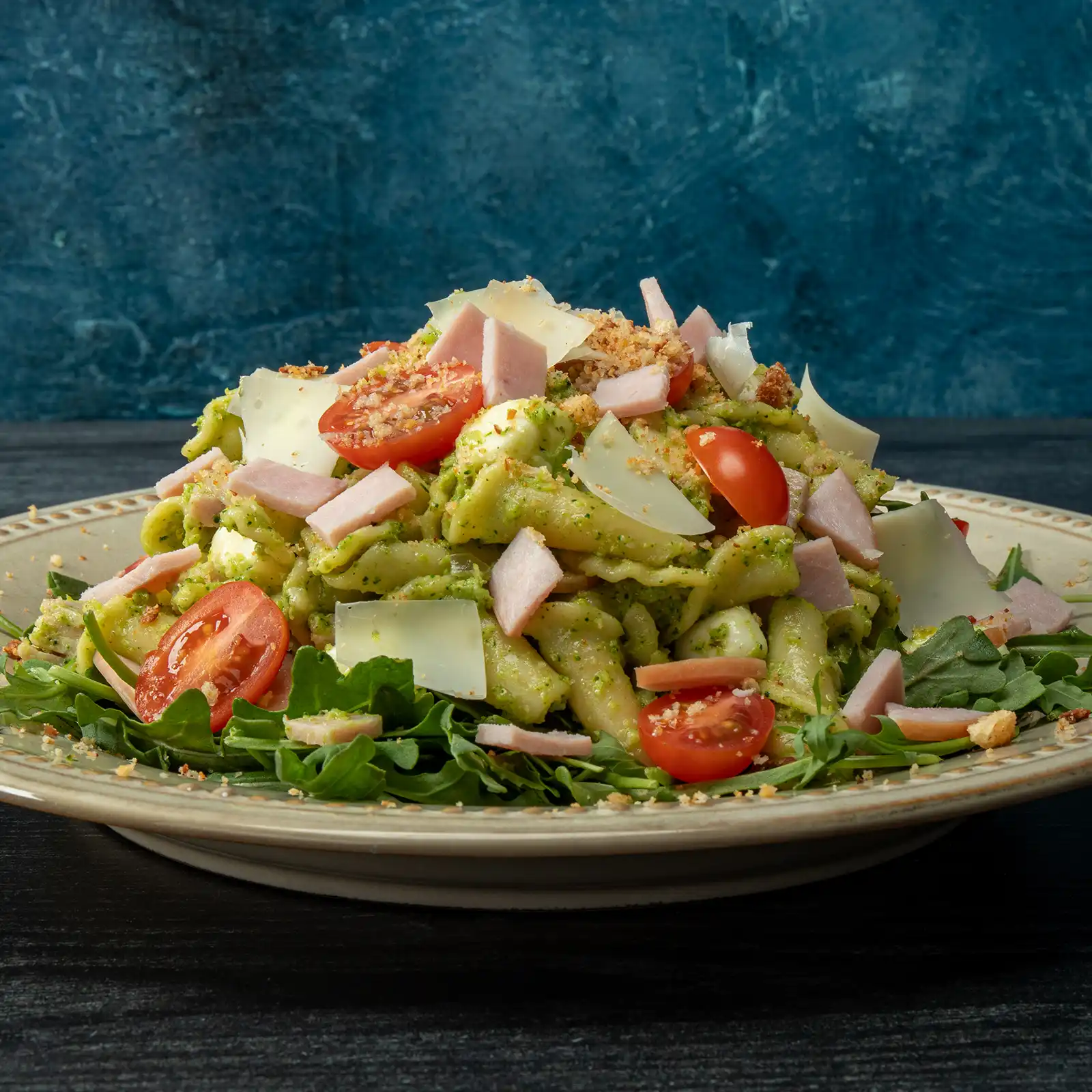 A mound of pasta tossed in bright green broccolini pesto, served over baby arugula and topped with diced Canadian bacon, halved cherry tomatoes, shaved Pecorino Romano, and toasted garlic breadcrumbs on a cream-rimmed plate against a deep blue background.