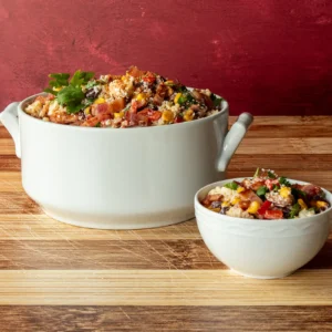 Street corn pasta salad with bacon served in a white handled soup pot and individual bowl, garnished with cilantro, Cotija cheese, and Tajin, on a bamboo surface with a red backdrop.