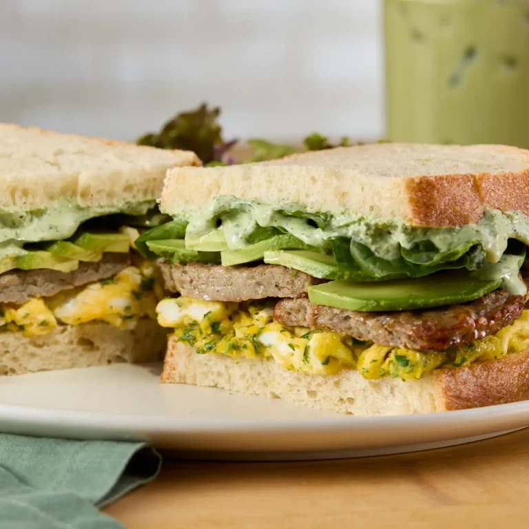 A turkey sausage breakfast sandwich on white bread, cut in half and stacked to show layers of herb-scrambled eggs, a Jones Dairy Farm turkey sausage patty, sliced avocado, mixed greens, and green herb sauce, served on a white plate with a teal napkin