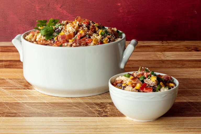 Street corn pasta salad with bacon served in a white handled soup pot and individual bowl, garnished with cilantro, Cotija cheese, and Tajin, on a bamboo surface with a red backdrop.
