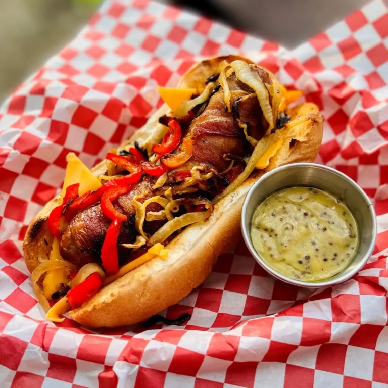 A bacon-wrapped bratwurst topped with grilled peppers, caramelized onions, and melted cheese, served on a toasted bun over red and white checkered paper, with a side of stone ground mustard vinaigrette.