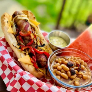 A bacon-wrapped bratwurst topped with grilled peppers, caramelized onions, and melted cheese, served on a toasted bun over red and white checkered paper, with sides of stone ground mustard vinaigrette and baked beans.