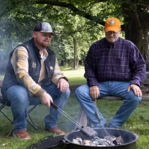 Will and Philip Jones sit around a campfire cooking pudgy pies in a cast iron pie iron, surrounded by lush green trees. Both wear Jones Dairy Farm branded hats and clothing, evoking the brand's multigenerational family heritage.