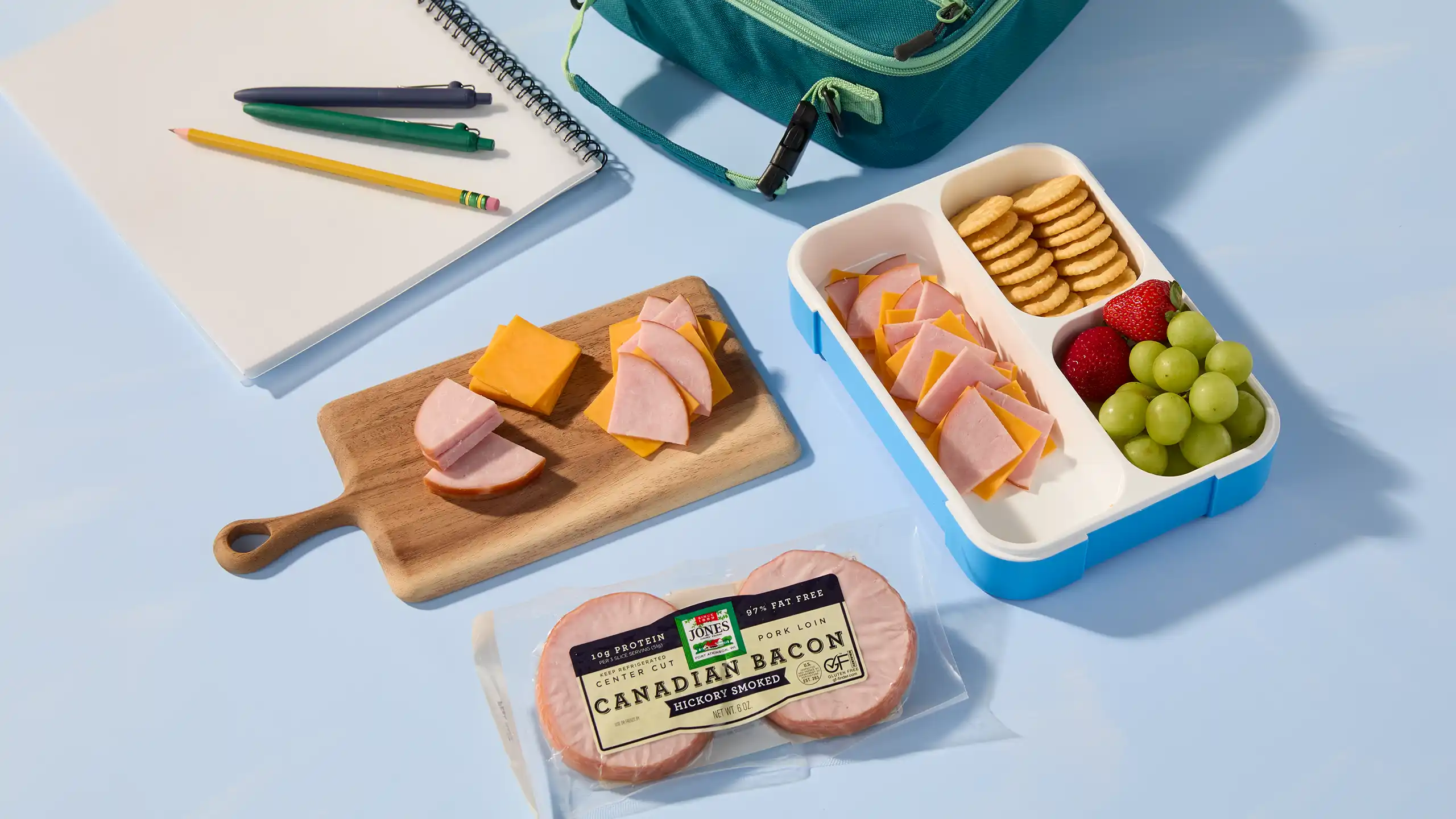 Jones Dairy Farm hickory smoked Canadian bacon next to a bento-style lunchbox with sliced Canadian bacon, cheddar cheese, crackers, strawberries, and grapes, styled with school supplies.