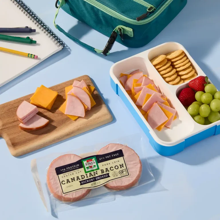 Back-to-school bento box with Jones Dairy Farm Canadian bacon and cheddar cheese cut into triangles, crackers, strawberries, and grapes, next to a teal lunch bag, notebook, and pencils.