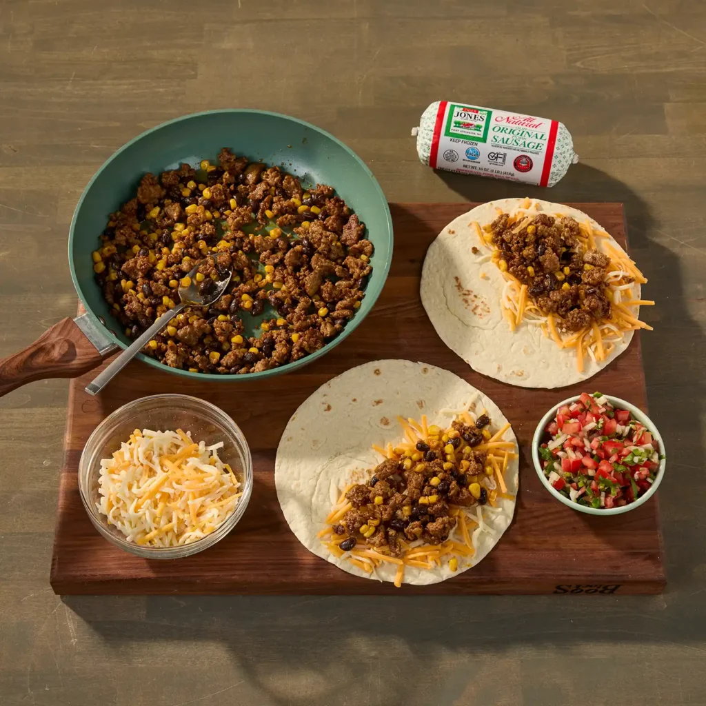 Overhead view of quesadilla assembly: a skillet of cooked sausage, corn, and black beans, flour tortillas topped with shredded cheese and filling, a bowl of pico de gallo, and a Jones All Natural Pork Sausage Roll on a cutting board.