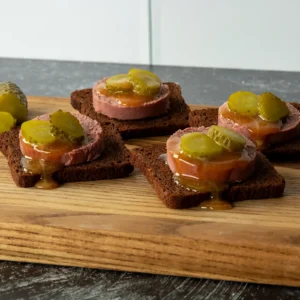 Close-up of Braunschweiger pumpernickel bites topped with dill pickle slices and mustard drizzle.