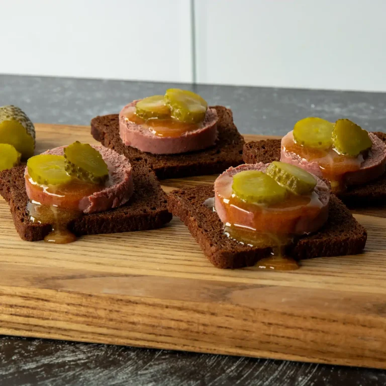 Close-up of Braunschweiger pumpernickel bites topped with dill pickle slices and mustard drizzle.