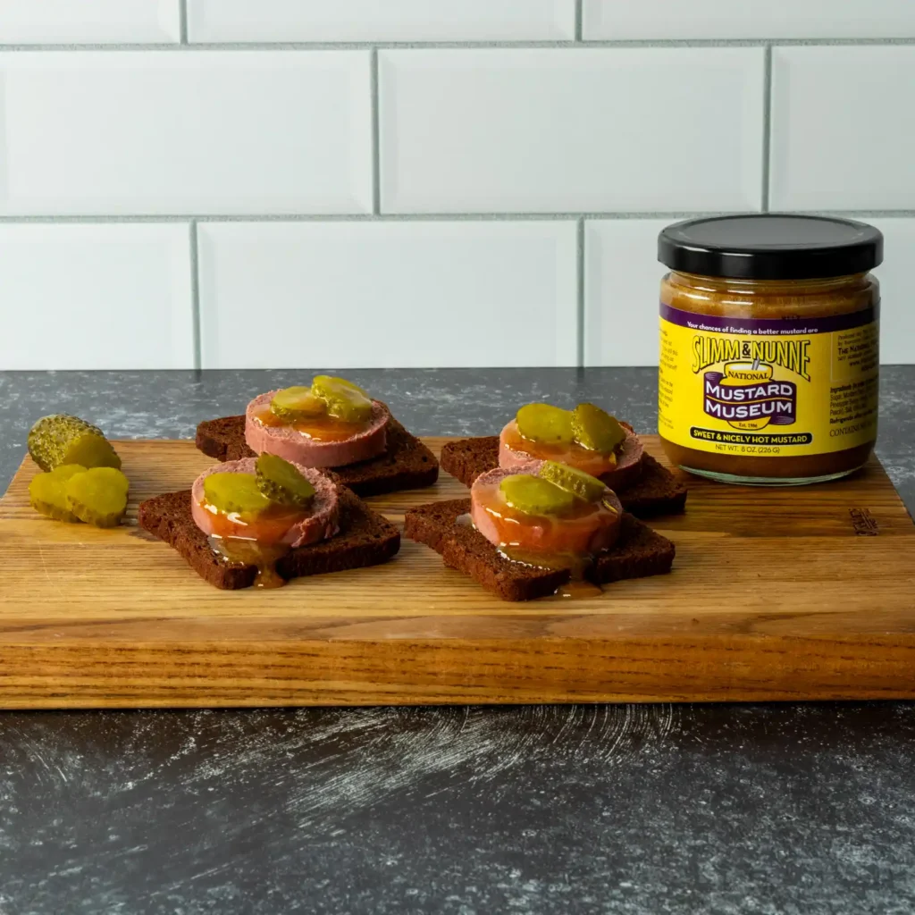 Braunschweiger pumpernickel bites topped with pickle, drizzled with mustard, next to a jar of Slimm & Nunne mustard.